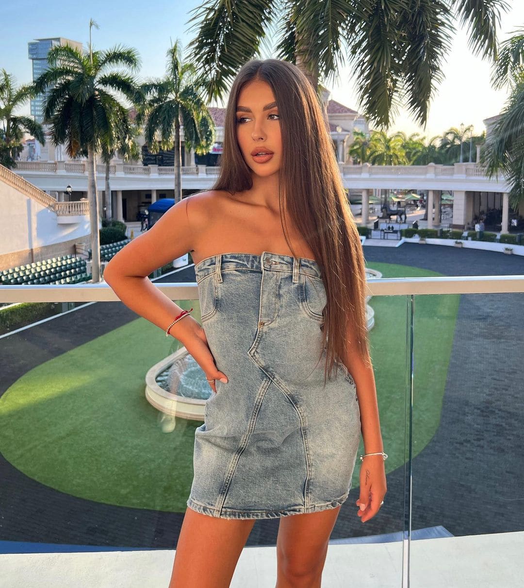 Lika in a strapless denim mini dress on a balcony with palm trees and a resort building behind her.