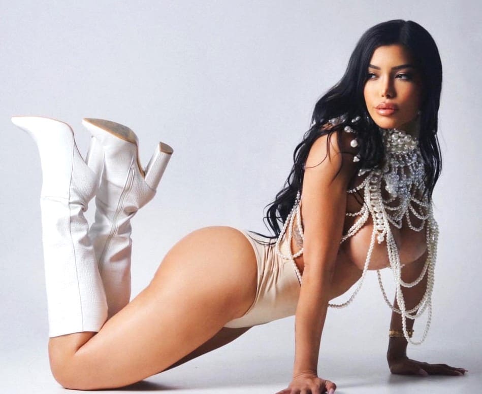 Mujer en cuatro patas en suelo blanco con bodysuit beige, botas blancas y top de perlas dejando torso expuesto.
