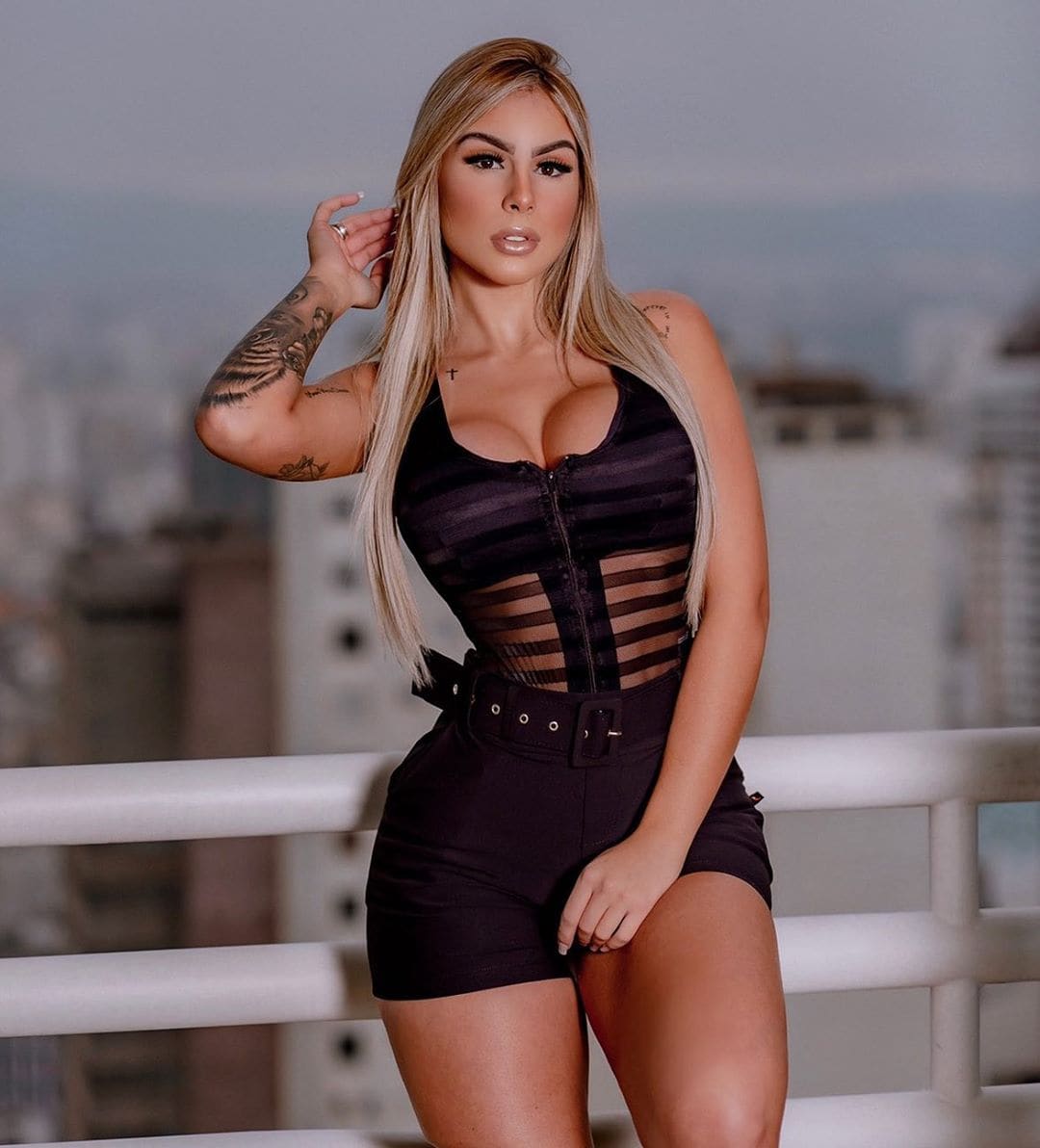 Mujer rubia con tatuajes en brazos en balcón, con top negro transparente rayado y shorts altos, mano en el pelo contra skyline borroso.