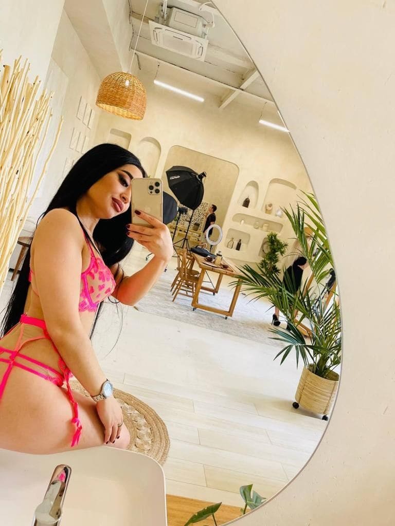 Donna con capelli scuri seduta su una superficie bianca, indossa un bikini rosa con top in pizzo e si fa un selfie in una stanza luminosa.
