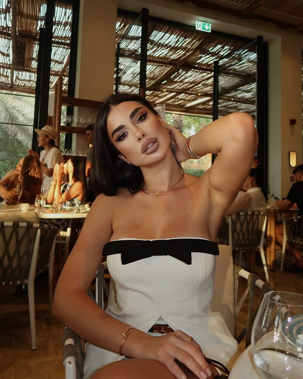 Femme assise dans un restaurant, robe blanche sans bretelles avec nœud noir, main près du cou.