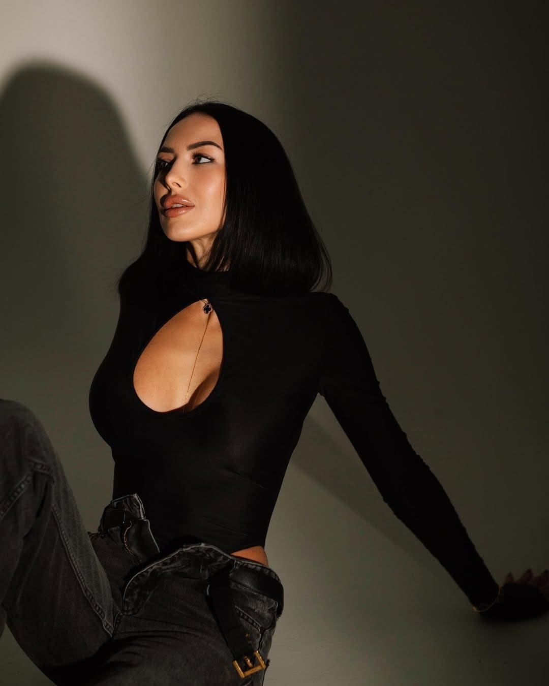 Mujer sentada en suelo gris con top negro de escote amplio y vaqueros oscuros.