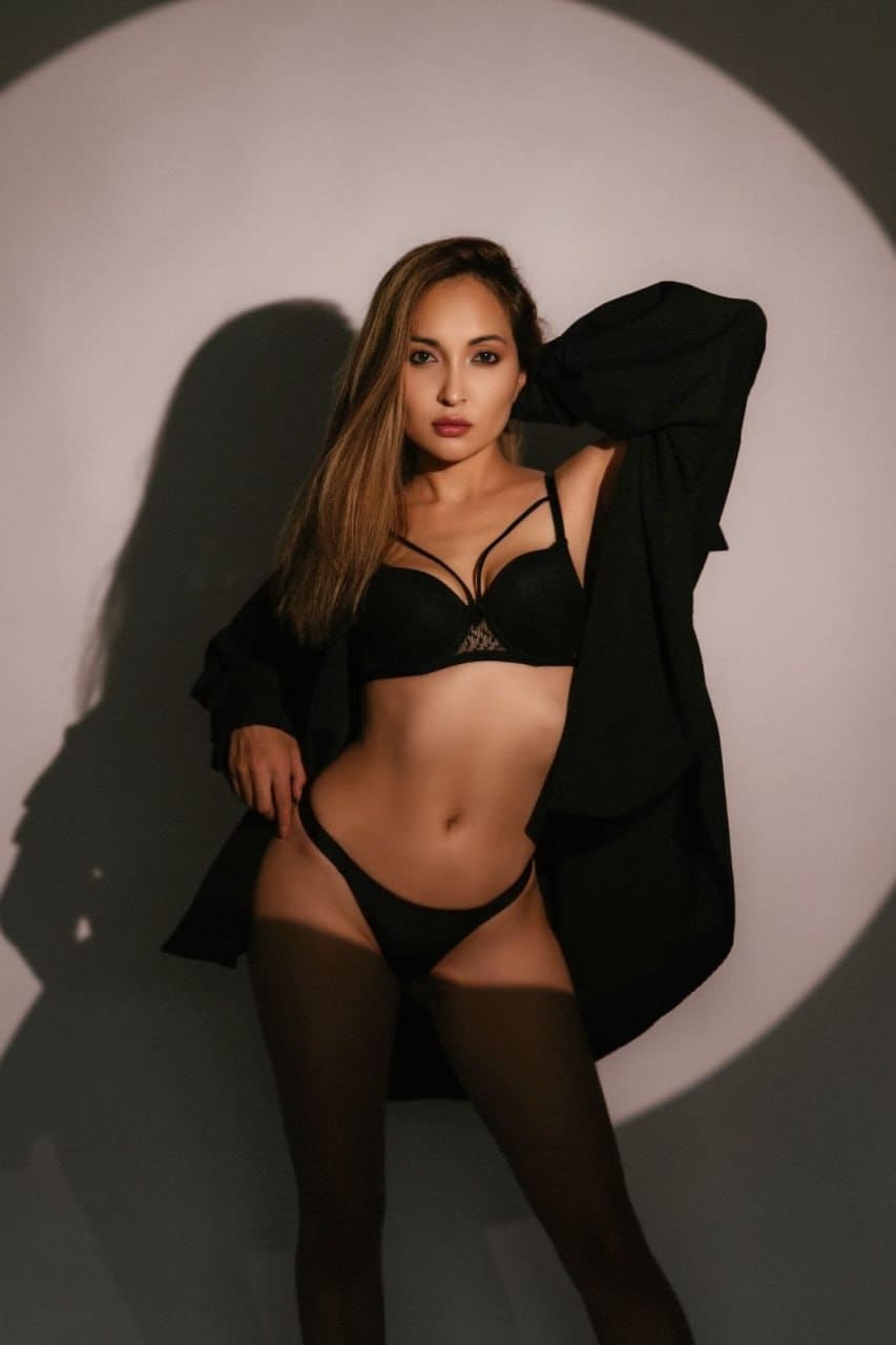 A woman in a black lace bra, briefs, tights, and open robe stands in spotlight with one hand behind her head.