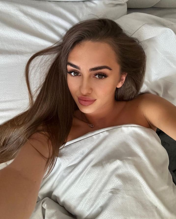 Mujer de cabello castaño largo tumbada en cama blanca con edredón estampado, hombros descubiertos mirando a cámara.
