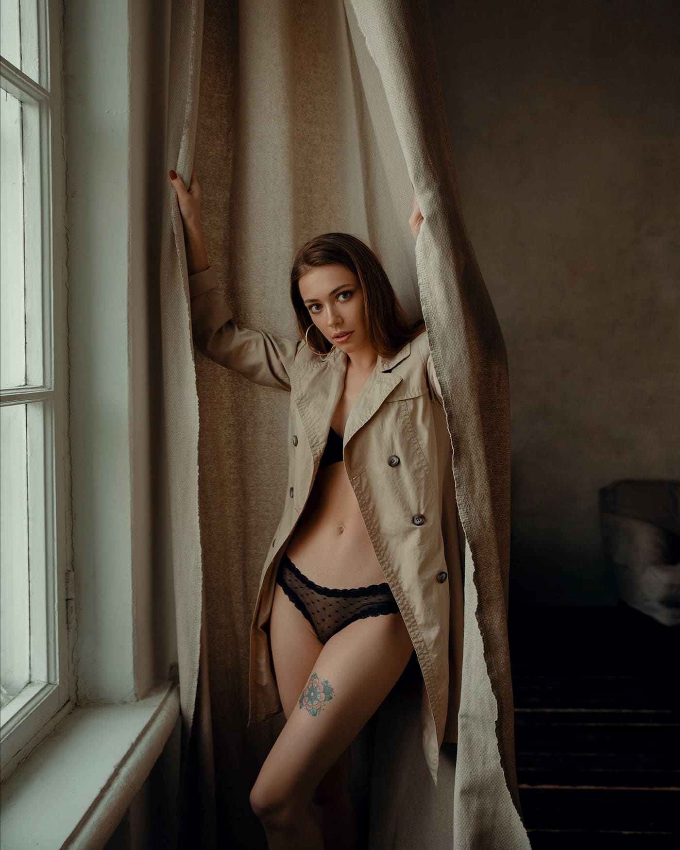 Mujer de cabello castaño entre cortinas beige, abrigo abierto sobre lencería negra, tatuaje floral en muslo izquierdo, habitación oscura.