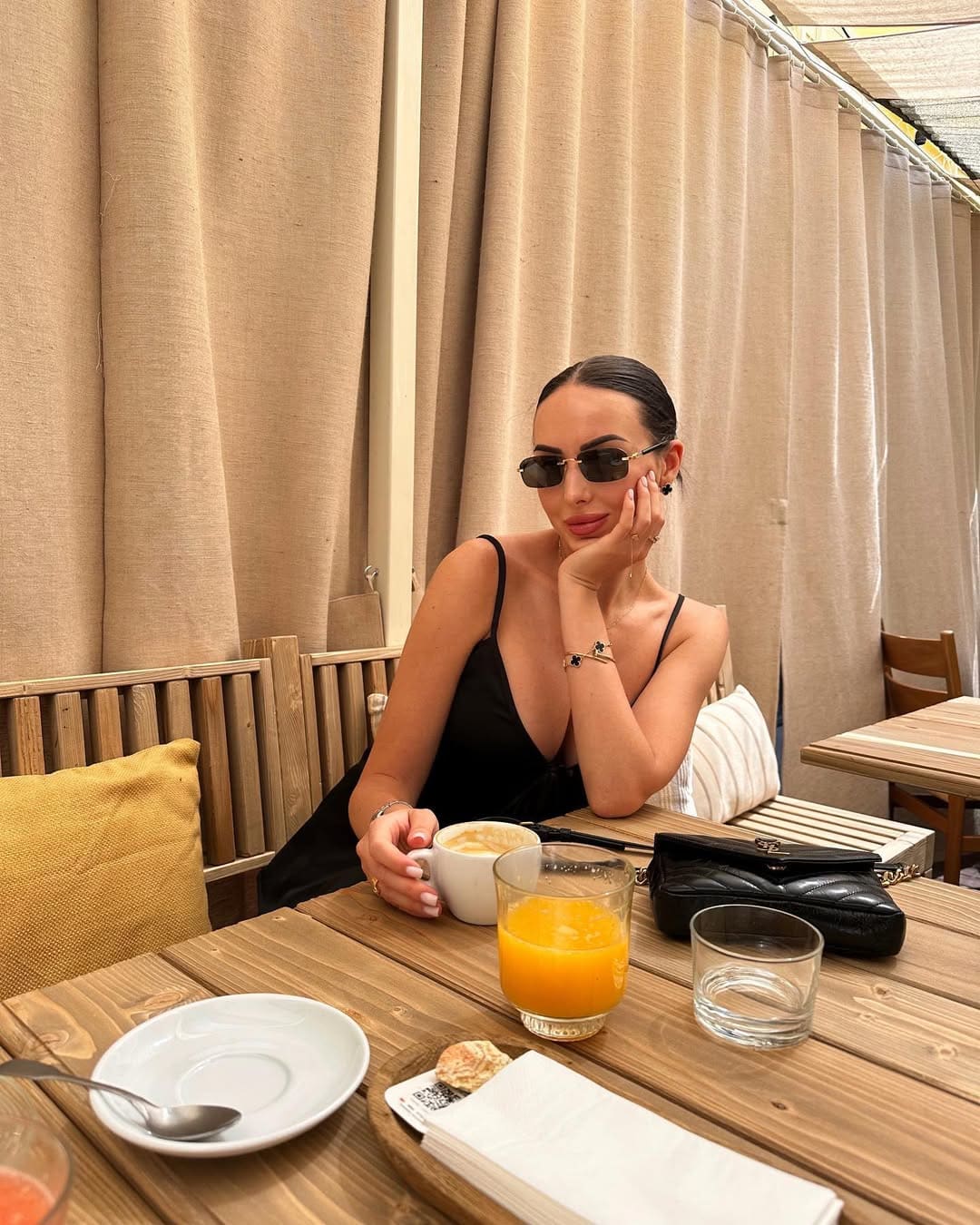 Mujer de top negro de tirantes y gafas sentada en mesa exterior con taza blanca.