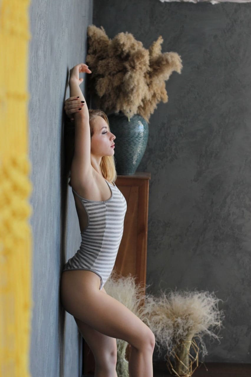Mujer rubia con bañador gris rayado y brazos levantados ante una pared gris, con jarrón y mueble al fondo.
