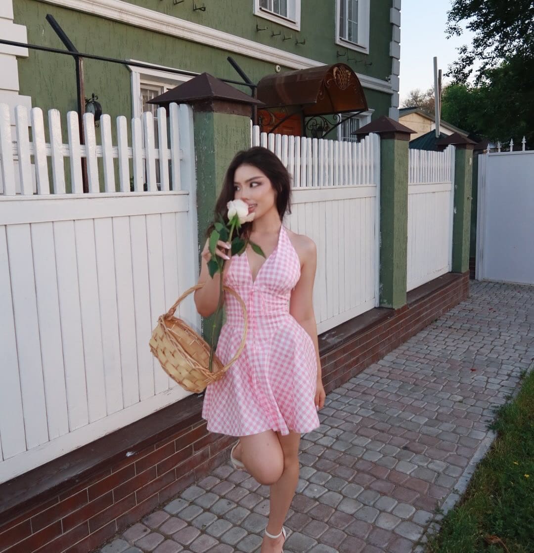 Mujer con pelo oscuro largo sobre un camino empedrado junto a una valla blanca, luciendo un vestido halter rosa y blanco a cuadros y sosteniendo una rosa.