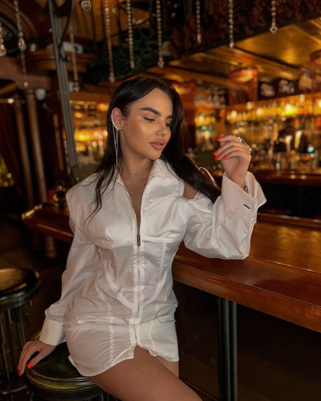 Mujer sentada en taburete de bar, chaqueta blanca abierta y mano cerca del rostro.