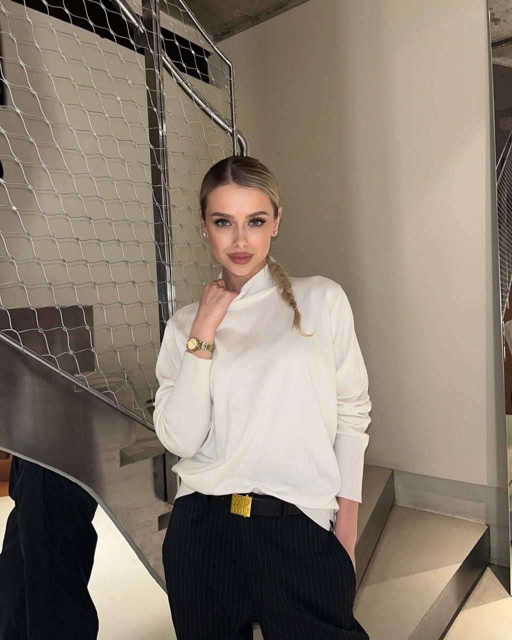 A woman with a side braid stands on a staircase in a white turtleneck and pinstripe trousers.