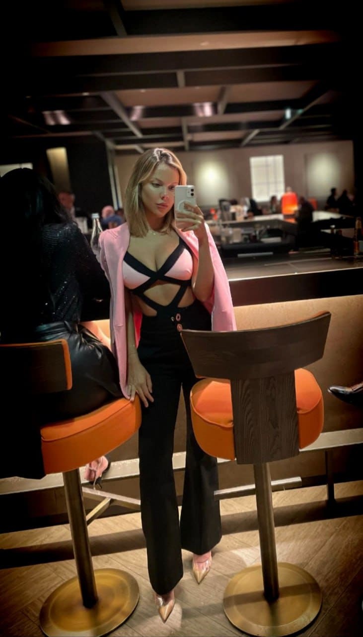 Selfie al espejo con blazer rosa y pantalón negro ancho, apoyada en una silla naranja en espacio tenue.