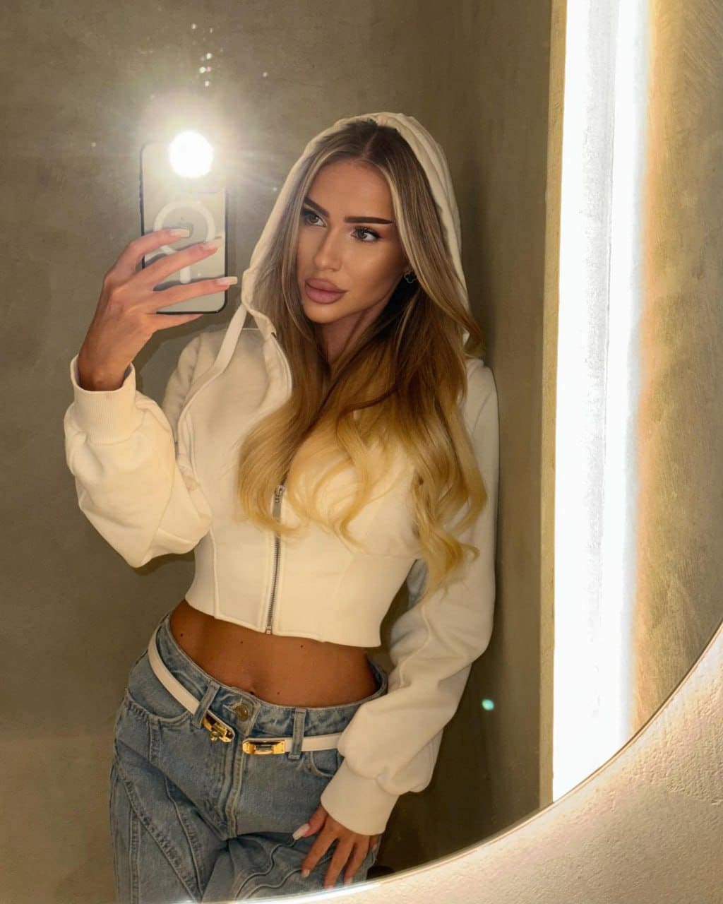 A woman takes a mirror selfie in a white cropped hoodie with hood up and light-wash jeans, facing a round mirror.