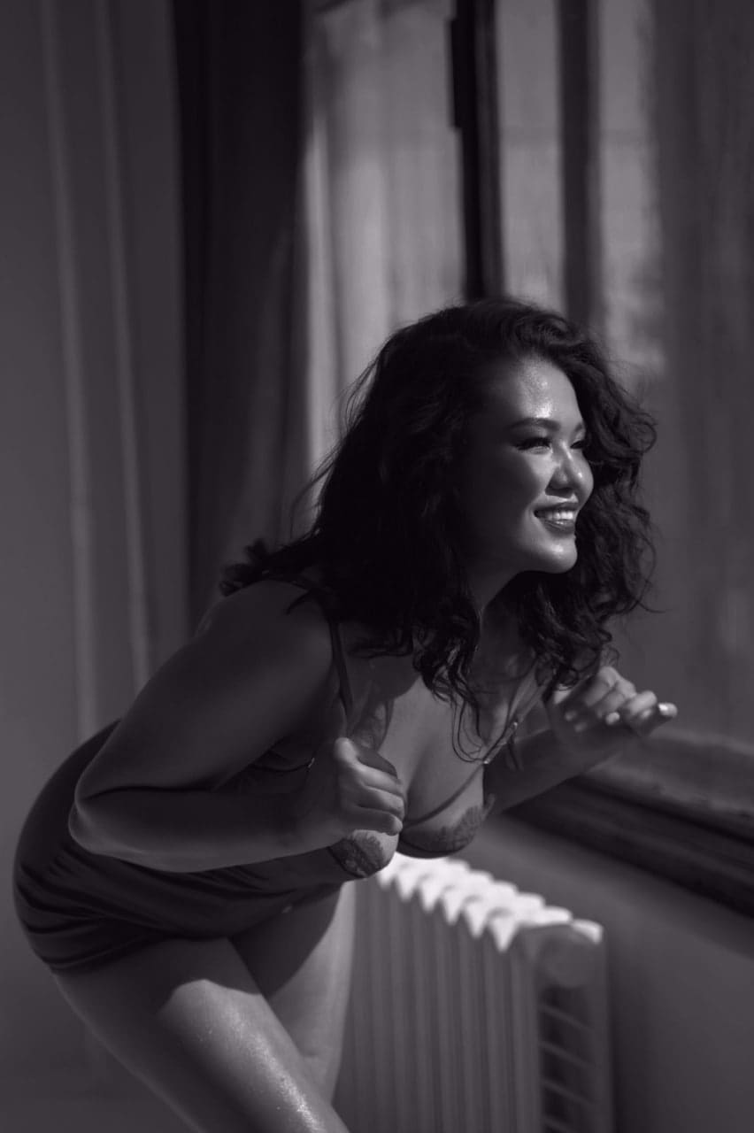 Mujer agachada junto a un radiador y ventana, vistiendo un vestido corto de tirantes.