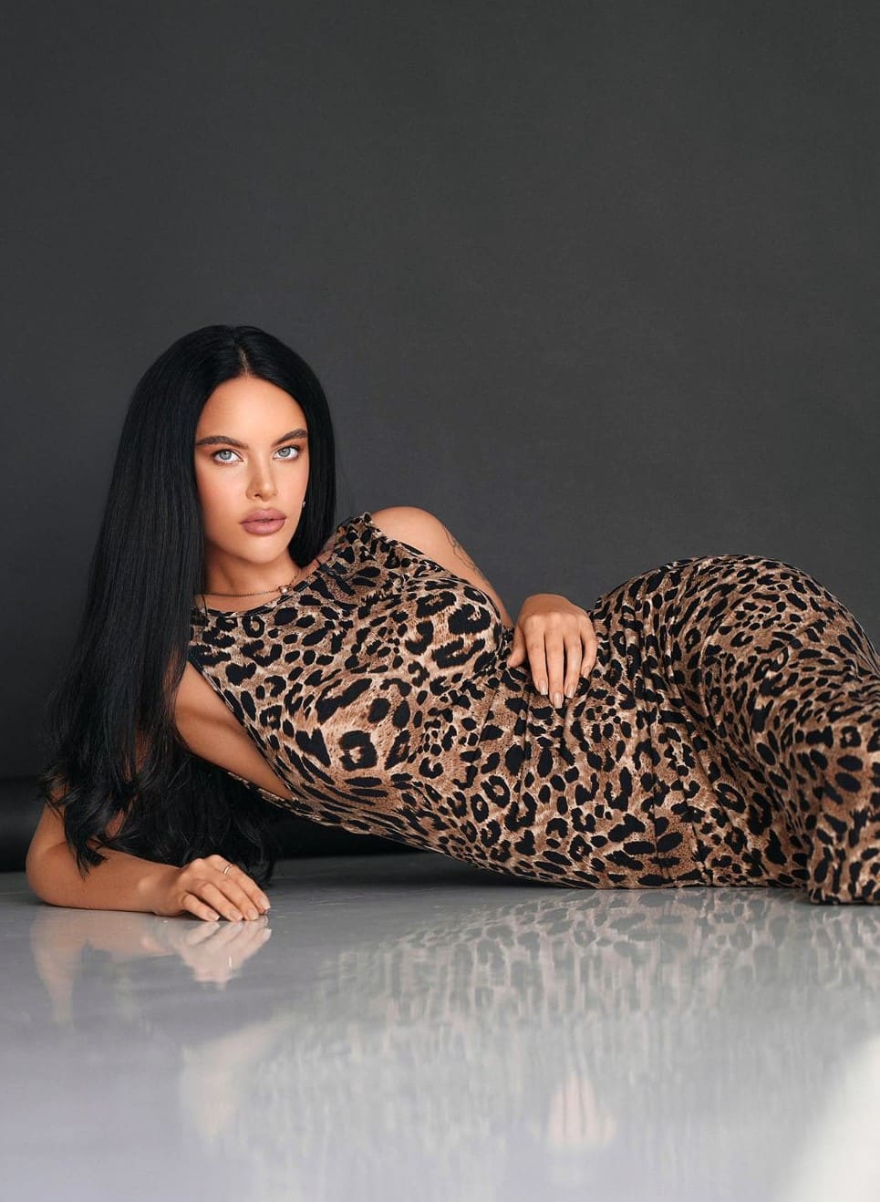 Mujer recostada sobre suelo gris reflectante con vestido leopardo ajustado y corte en el hombro, mirando a cámara.