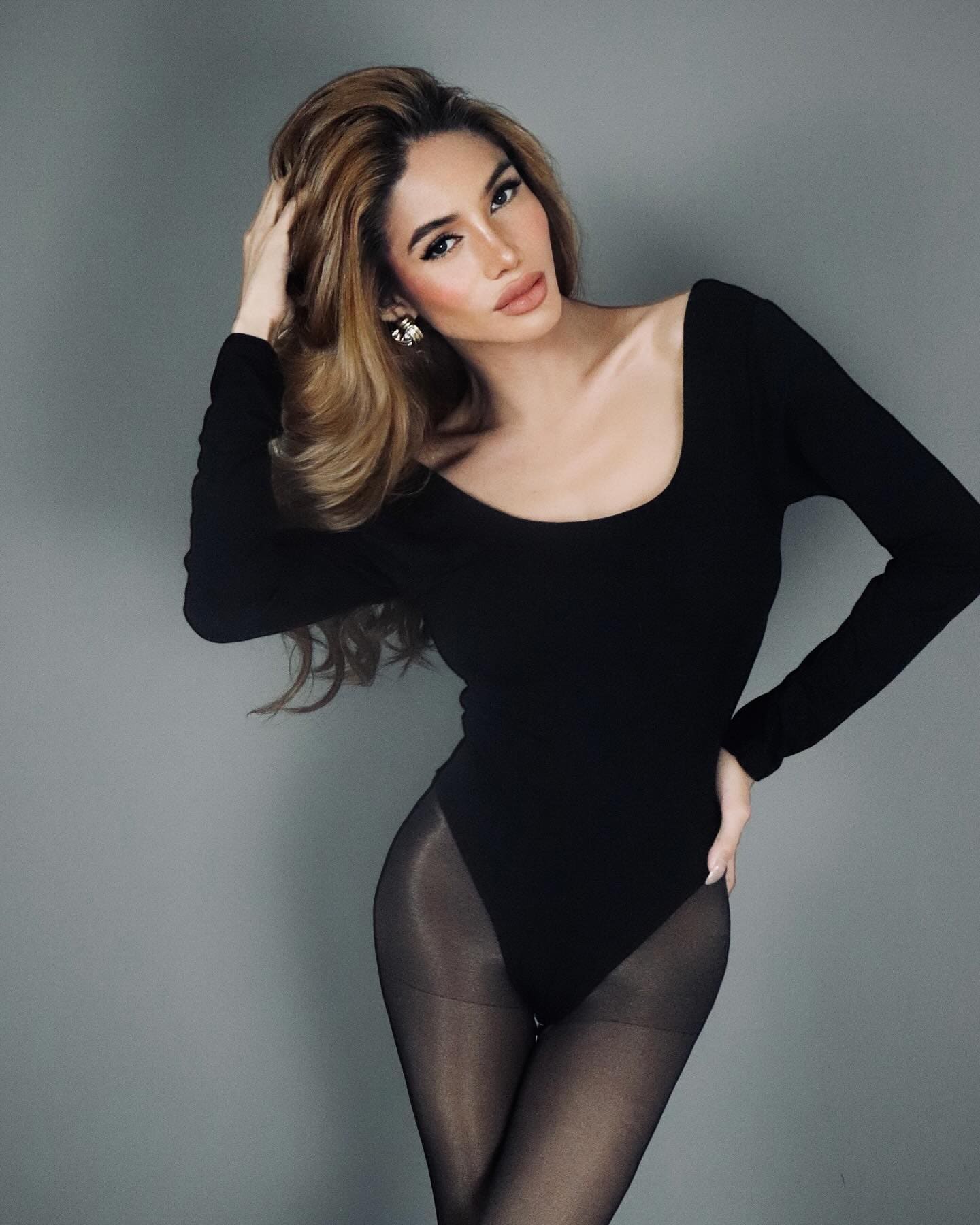 Nancy in a black bodysuit and tights stands against a gray backdrop, one hand raised to her hair.