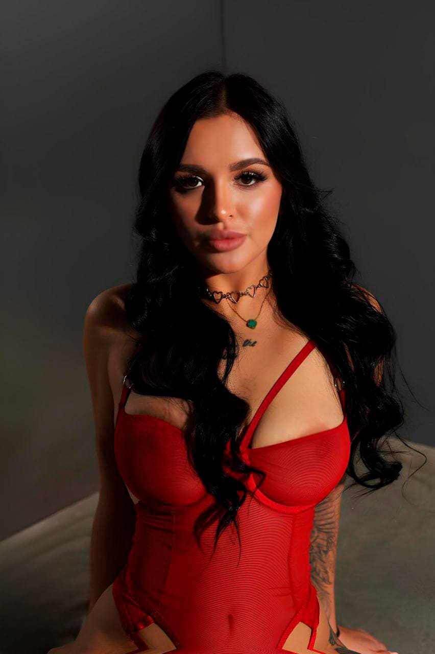 Alsu con cabello oscuro rizado viste un bodysuit rojo de tirantes finos y collares, sentada frente a cámara en estudio.