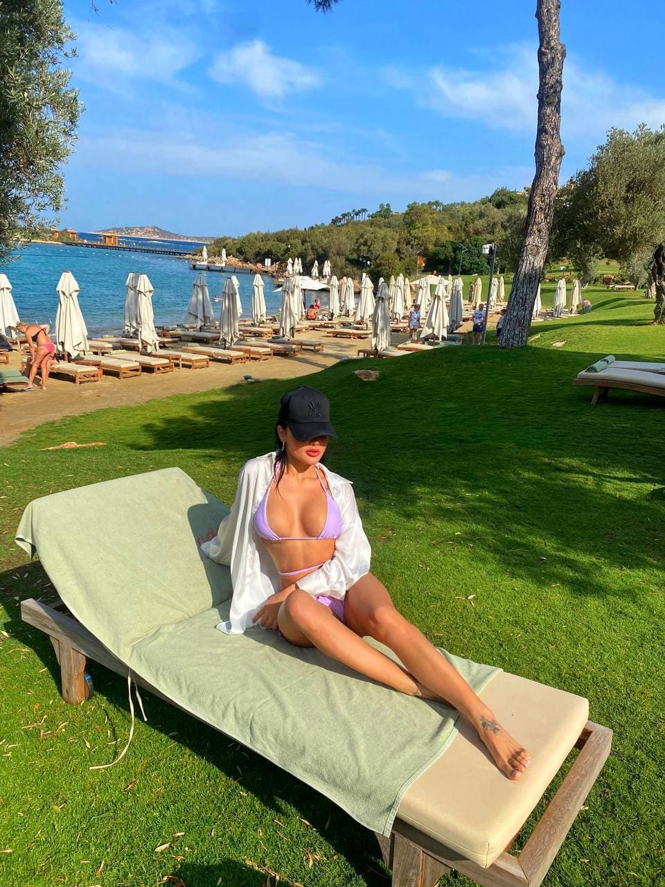 Mujer con gorra negra y camisa blanca abierta sobre bikini morada tumbada en una hamaca de césped con sombrillas blancas al fondo.