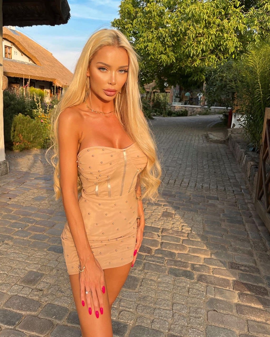 Anya con vestido beige sin tirantes y detalles de tachuelas posa en un camino empedrado frente a una choza.