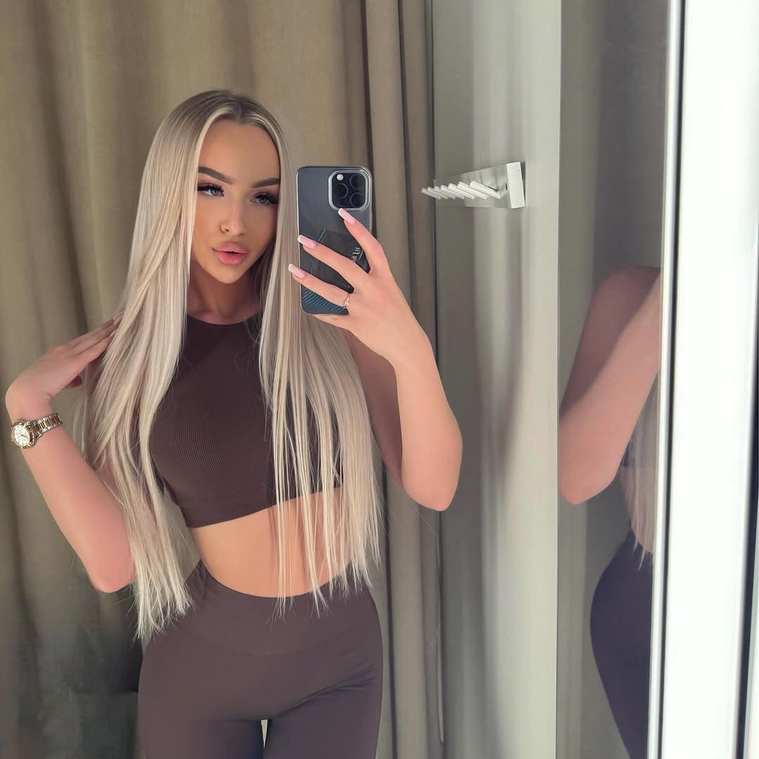 Svetlana takes a mirror selfie in a brown ribbed crop top and leggings, touching her hair near beige curtains.