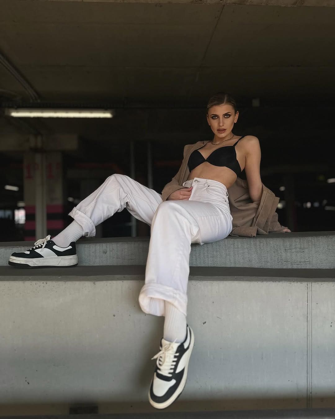 Mujer rubia sentada en un banco interior con poca luz, piernas cruzadas y chaqueta beige al cuello.