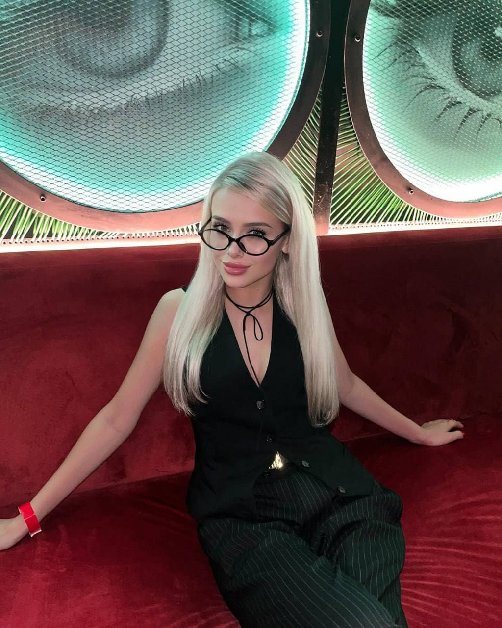 A woman in glasses sits on a red couch in a black V-neck top and dark pinstripe trousers.