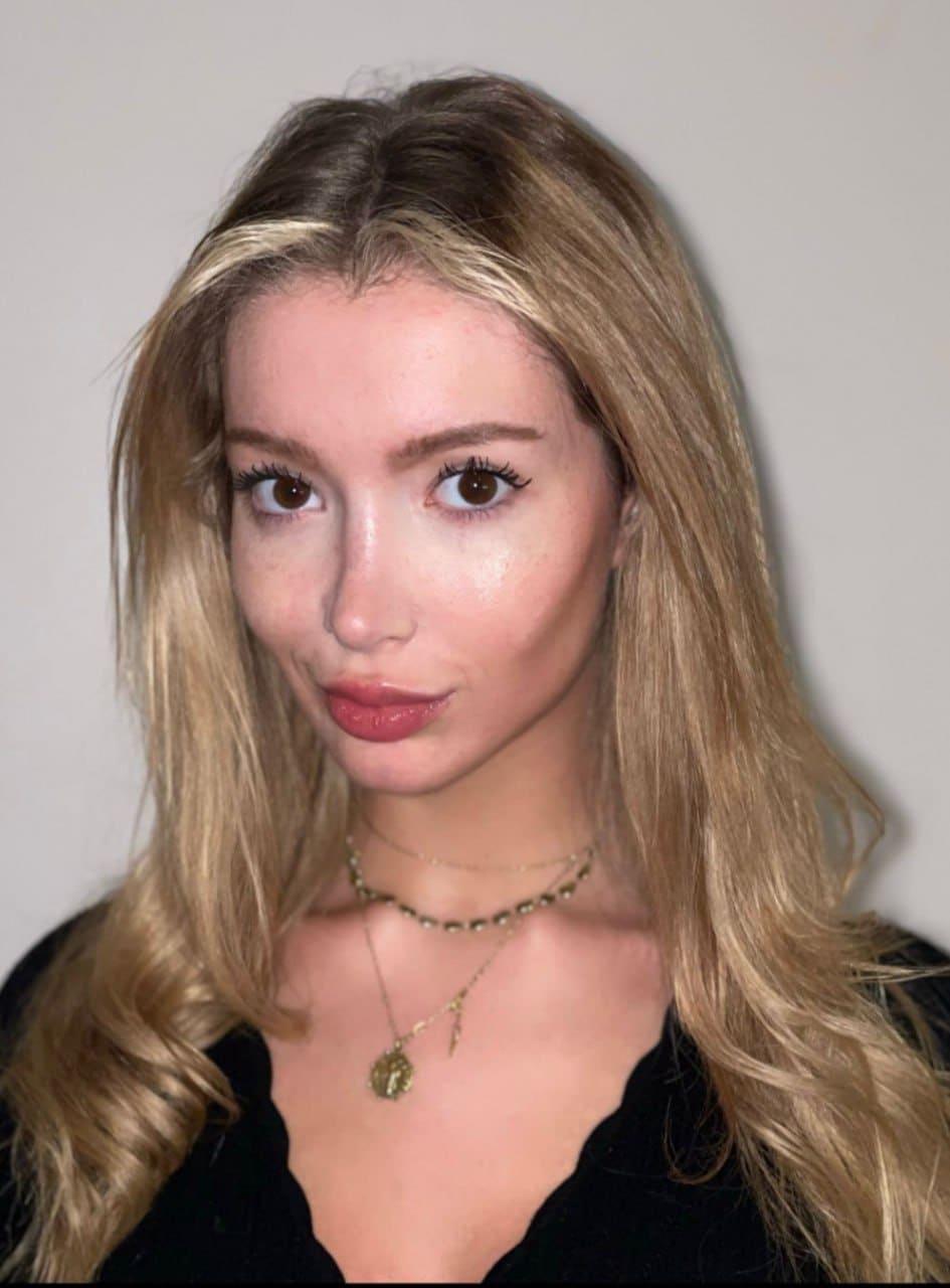 Clara in a black low-neck top and gold necklaces against a plain light wall.