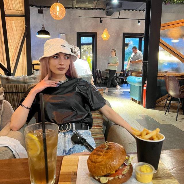 Alice con cabello rosa sentada en restaurante con sombrero claro, top negro y shorts vaqueros, frente a una hamburguesa.