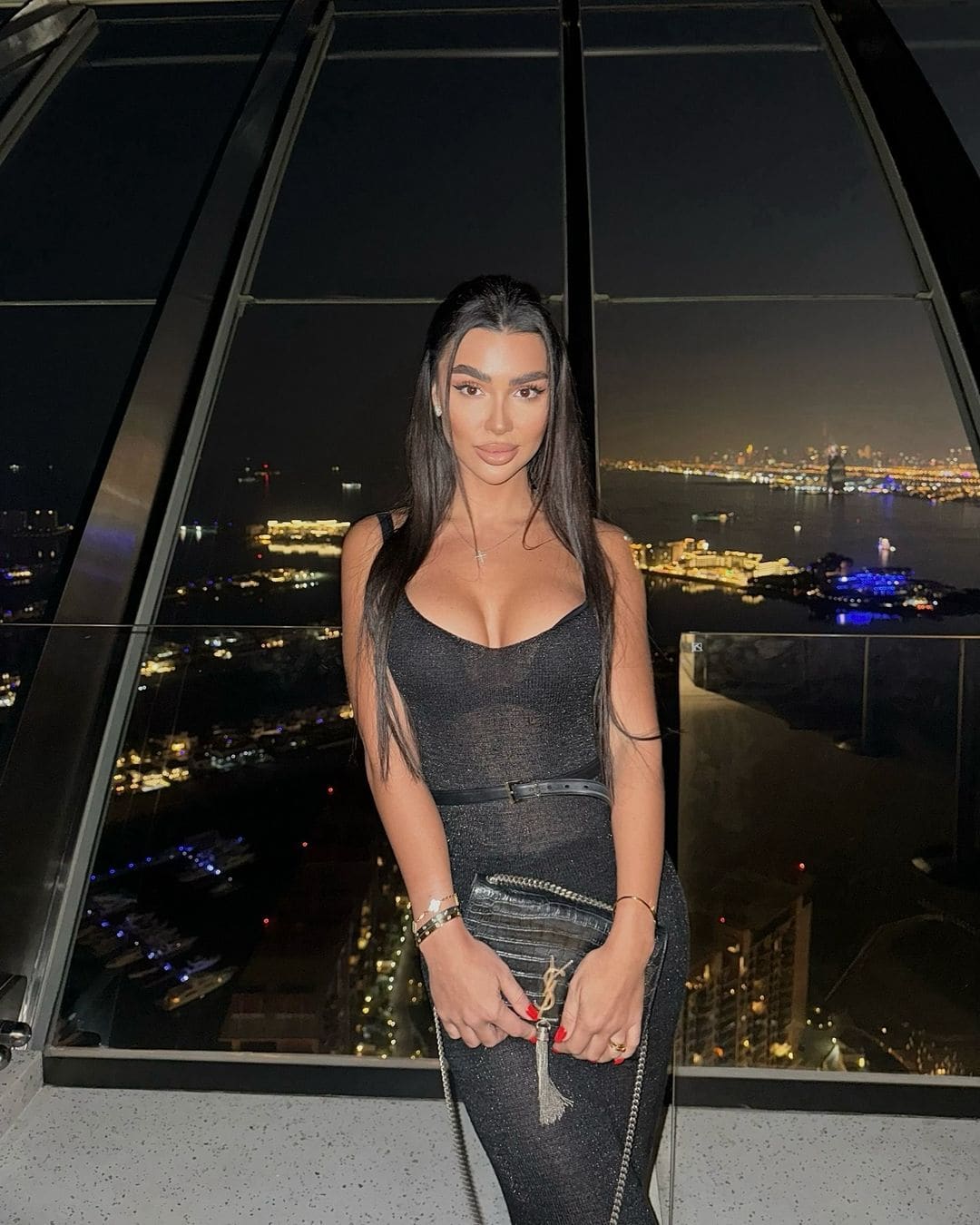 Mujer con vestido oscuro ajustado y cinturón se apoya junto a ventana nocturna urbana, sosteniendo bolso pequeño con correa de cadena.