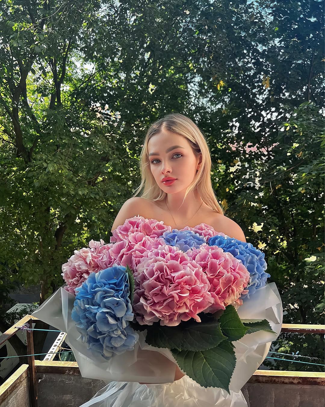 Mujer rubia con flequillo recto sujetando un ramo de hidrangéas rosas y azules, con top sin tirantes claro ante follaje verde.