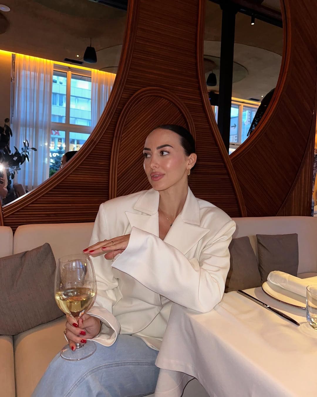 Mujer con blazer blanco y vaqueros sentada en sofá beige de restaurante bebiendo vino.