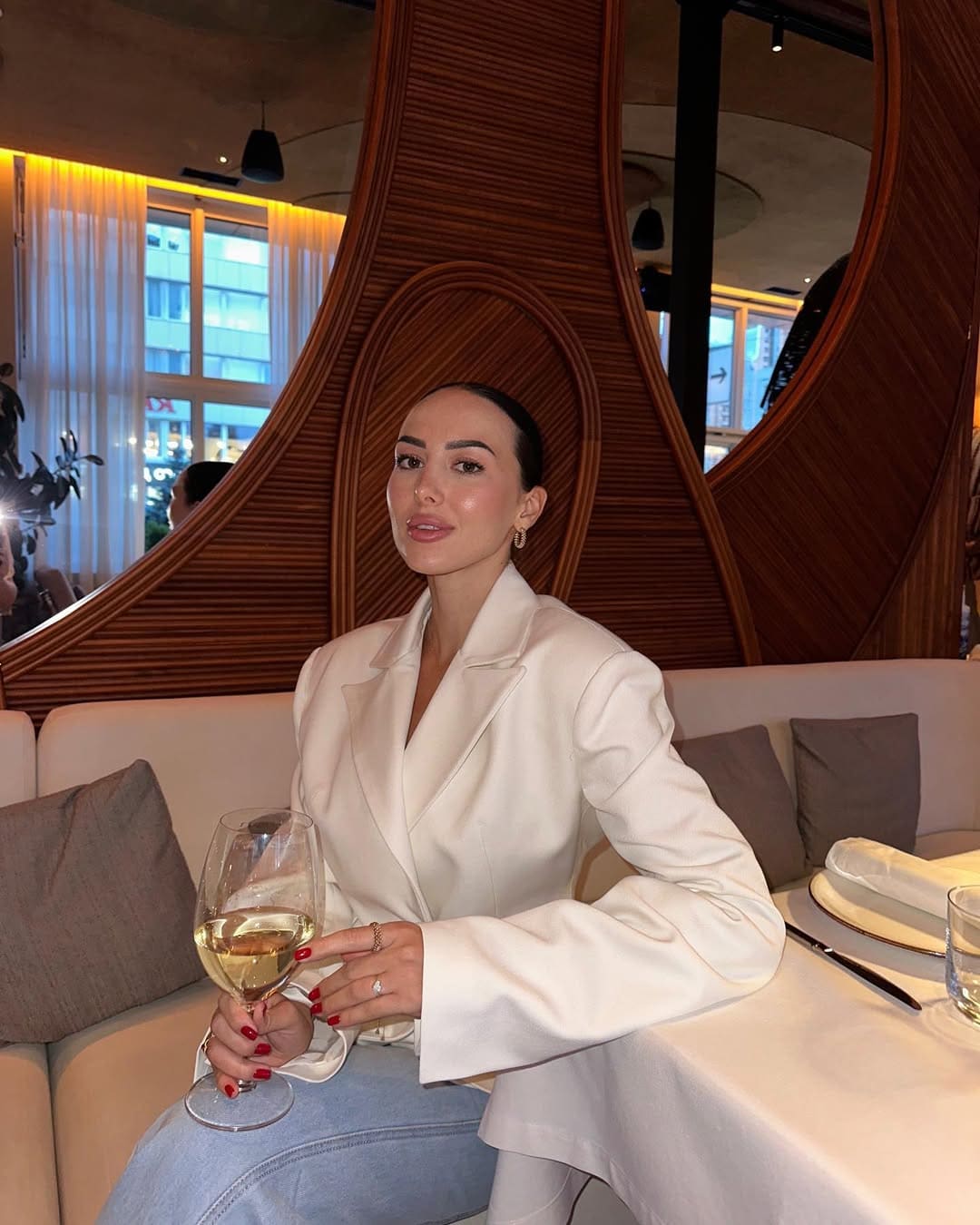 Juliette sitzt an einem Restauranttisch in weißem Blazer und Jeans, hält ein Weinglas vor hölzerner Trennwand.