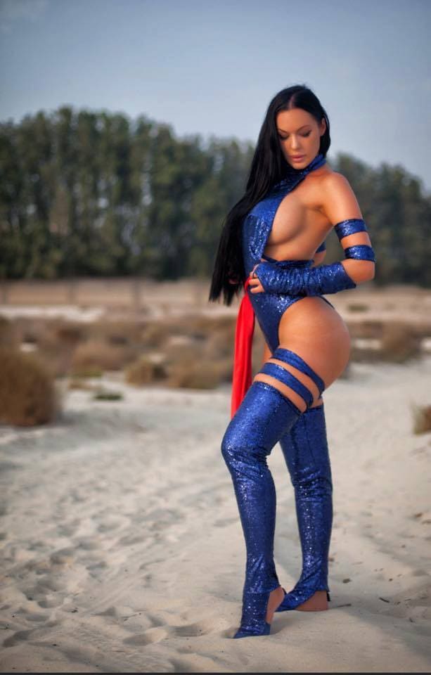 Mujer en playa arenosa, con bodysuit azul brillante de escote profundo, botas altas a juego y fajín rojo, árboles al fondo.