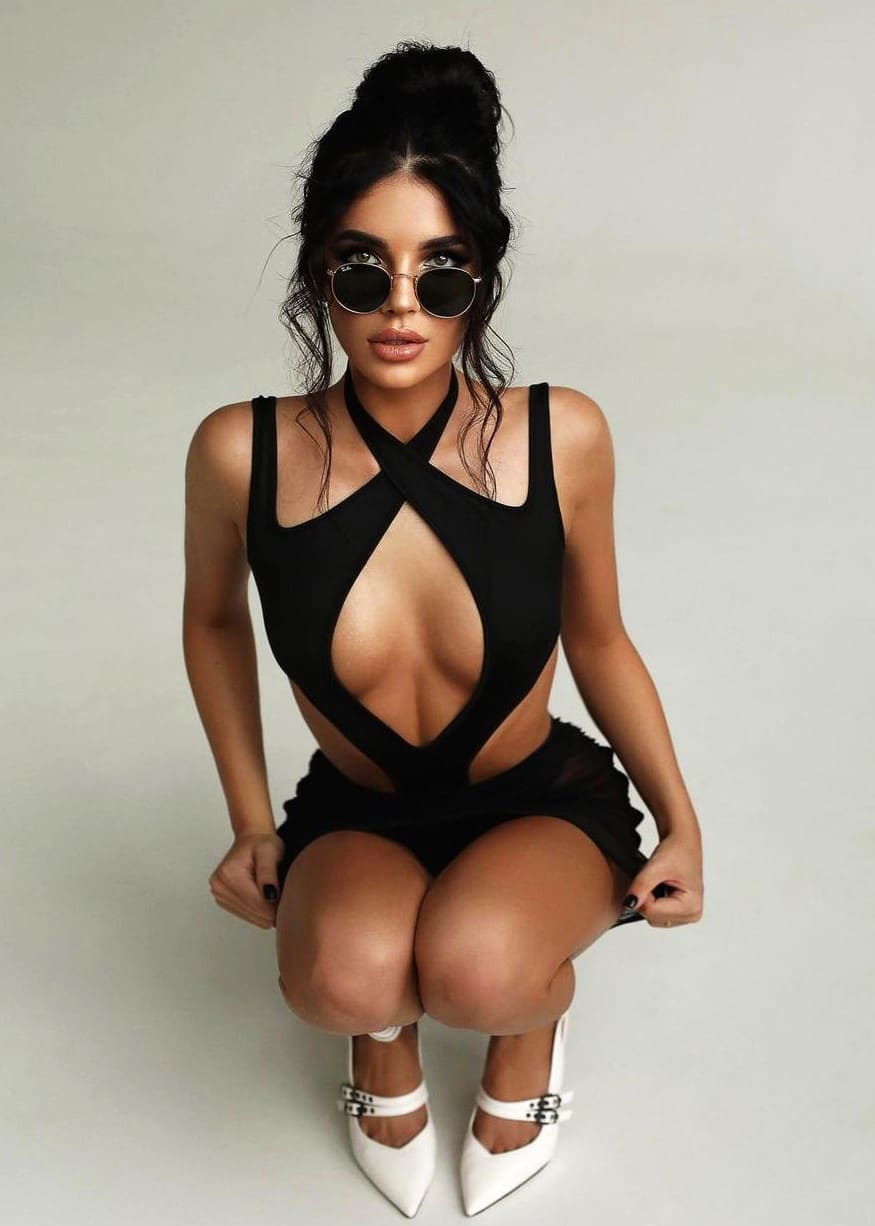 Donna con chignon alto in abito nero con tagli, occhiali rotondi e scarpe bianche a punta, accovacciata.