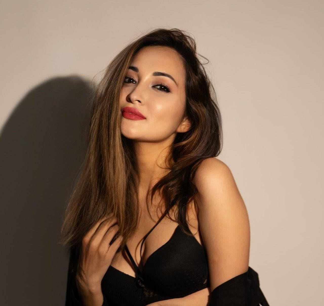 Chest-up view of a woman in a black bra and off-shoulder garment against a light wall with a shadow to the left.
