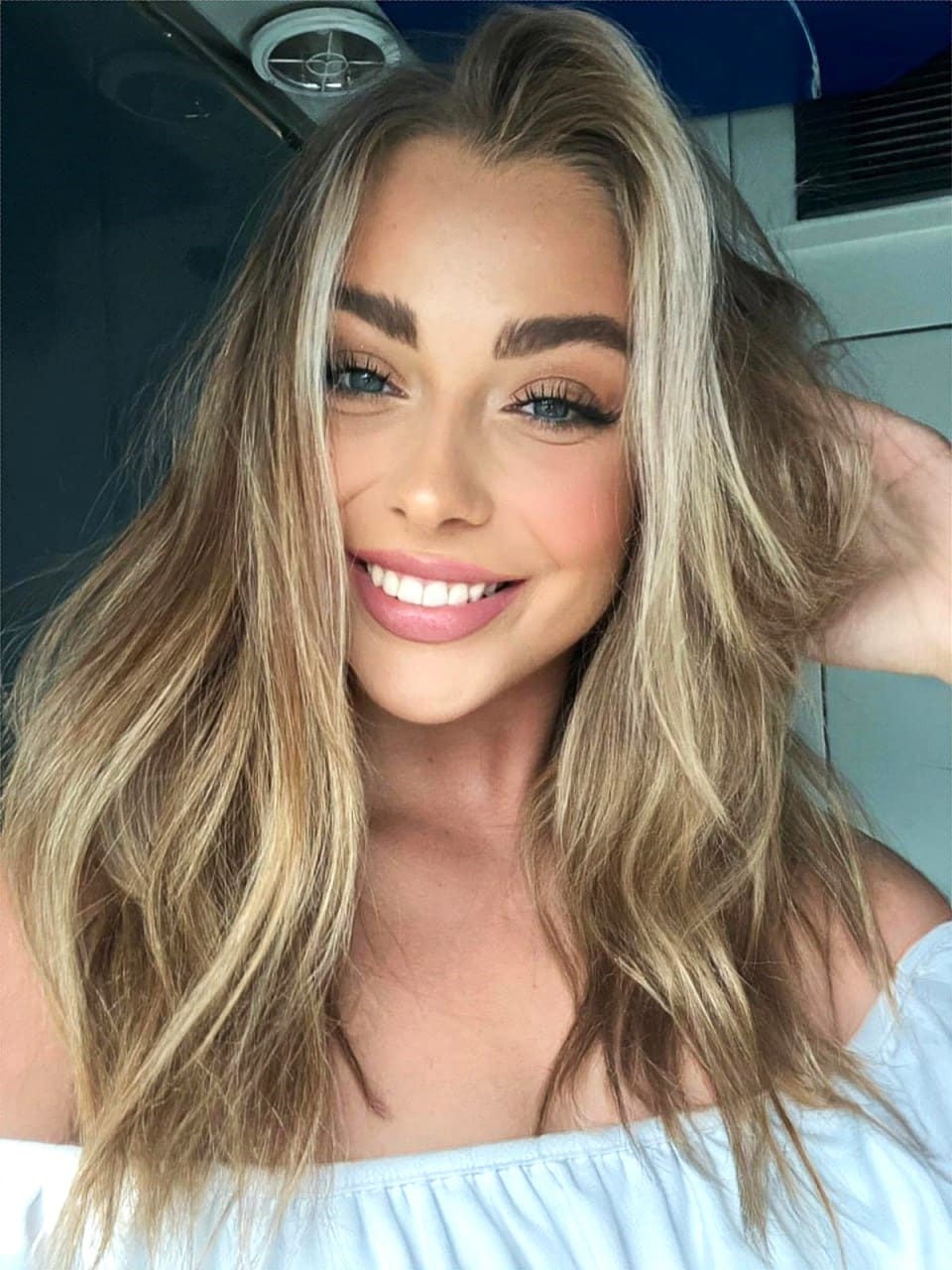 Mujer rubia sonriendo con blusa azul claro al descubierto del hombro, tocándose el pelo en interior iluminado.