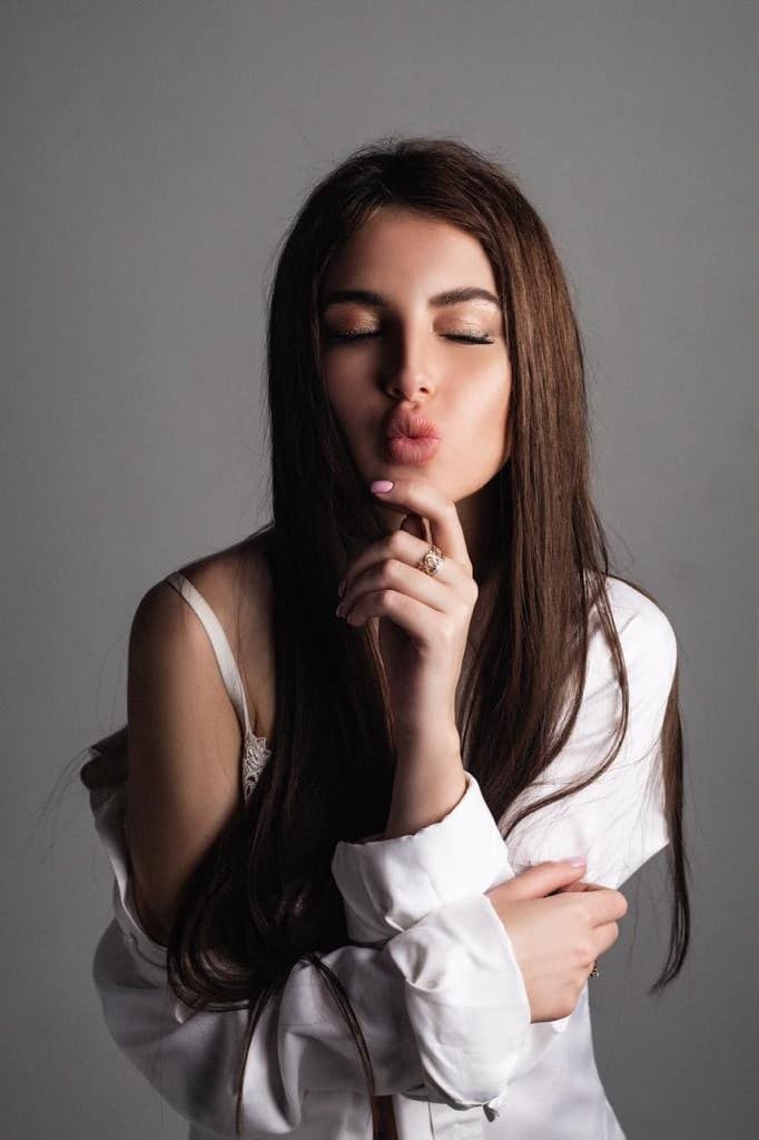 A woman in a white shirt slipping off one shoulder poses with eyes closed and finger on chin against a plain gray background.