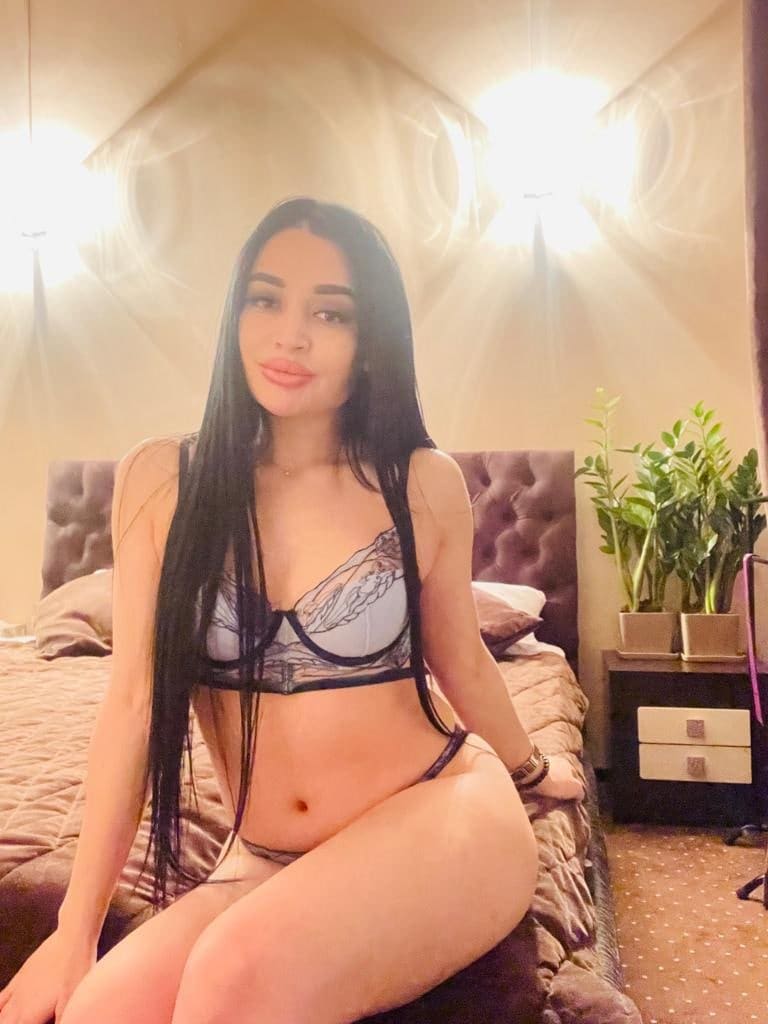 Donna con capelli scuri seduta su un letto, indossa un set di lingerie in pizzo a due pezzi, con testiera imbottita e piante sullo sfondo.