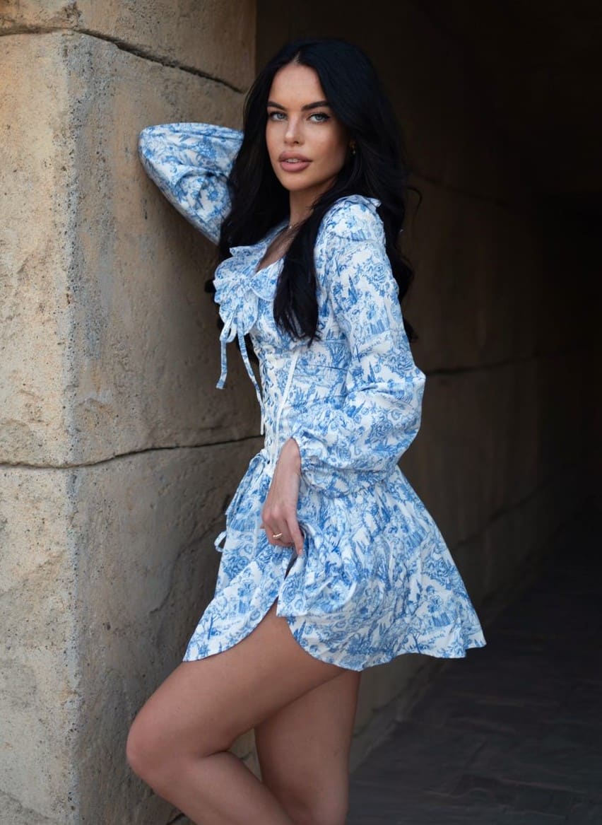 Mujer apoyada en muro de piedra con vestido azul y blanco estampado de mangas largas y cuello fruncido.