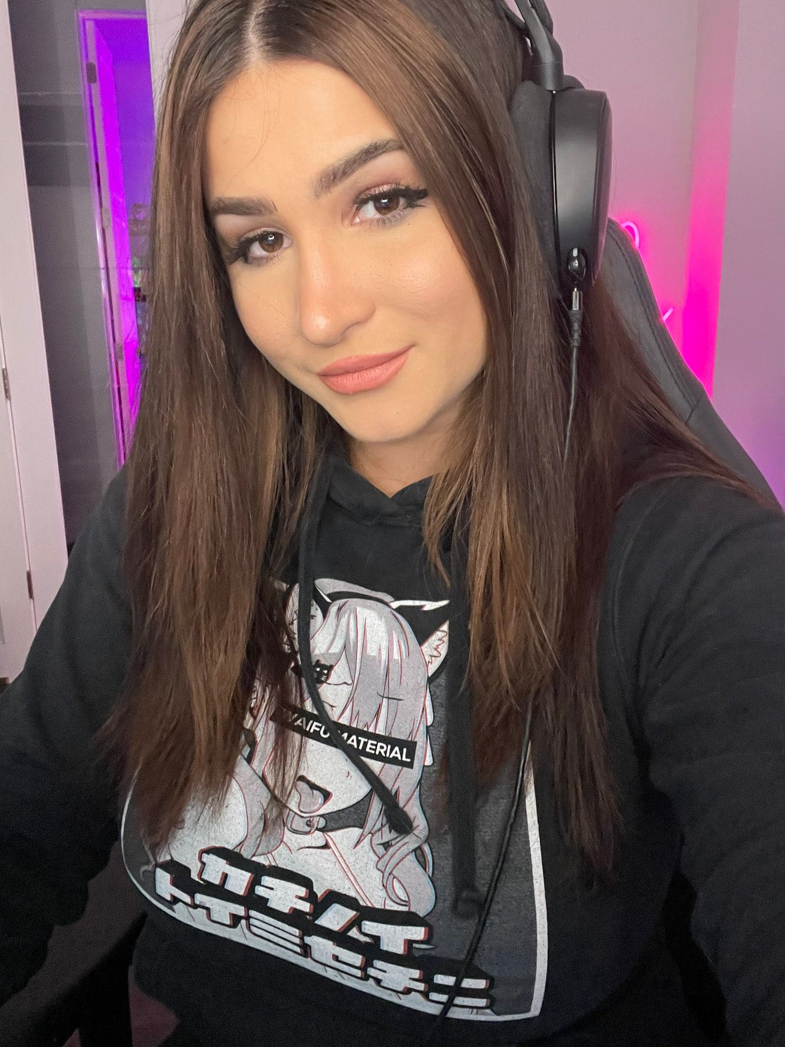 Joven con sudadera negra gráfica y auriculares, sentada en silla gamer bajo luz neón morada.