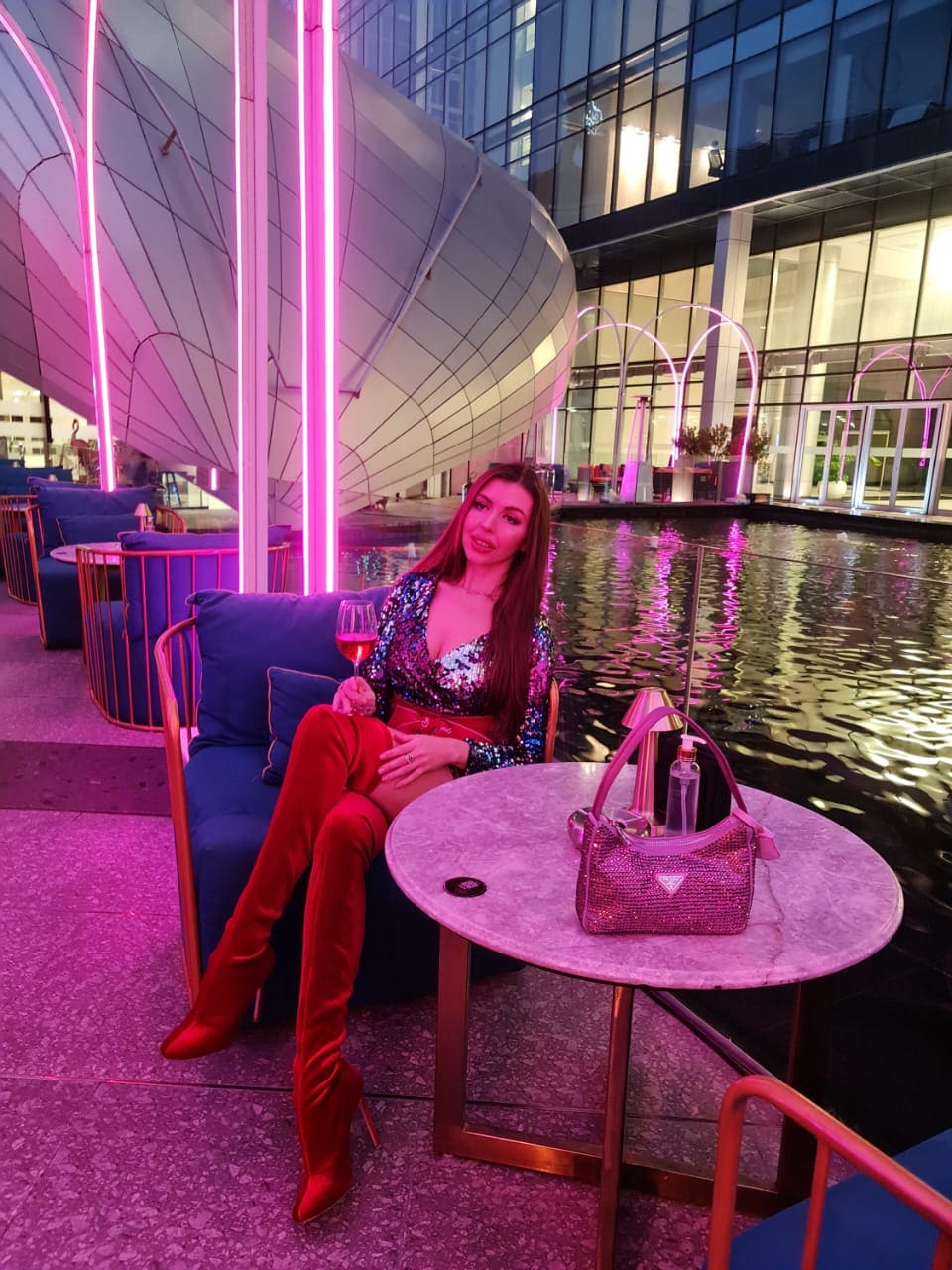 Mujer sentada en silla azul junto a piscina de noche, con top de lentejuelas, botas rojas altas y copa de vino en mano.
