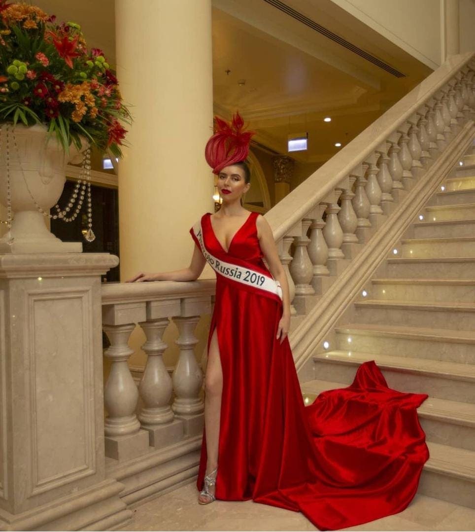 Mujer con peinado alto y tocado de plumas rojas en escalera de mármol, con vestido rojo de satén y mano en el barandal.