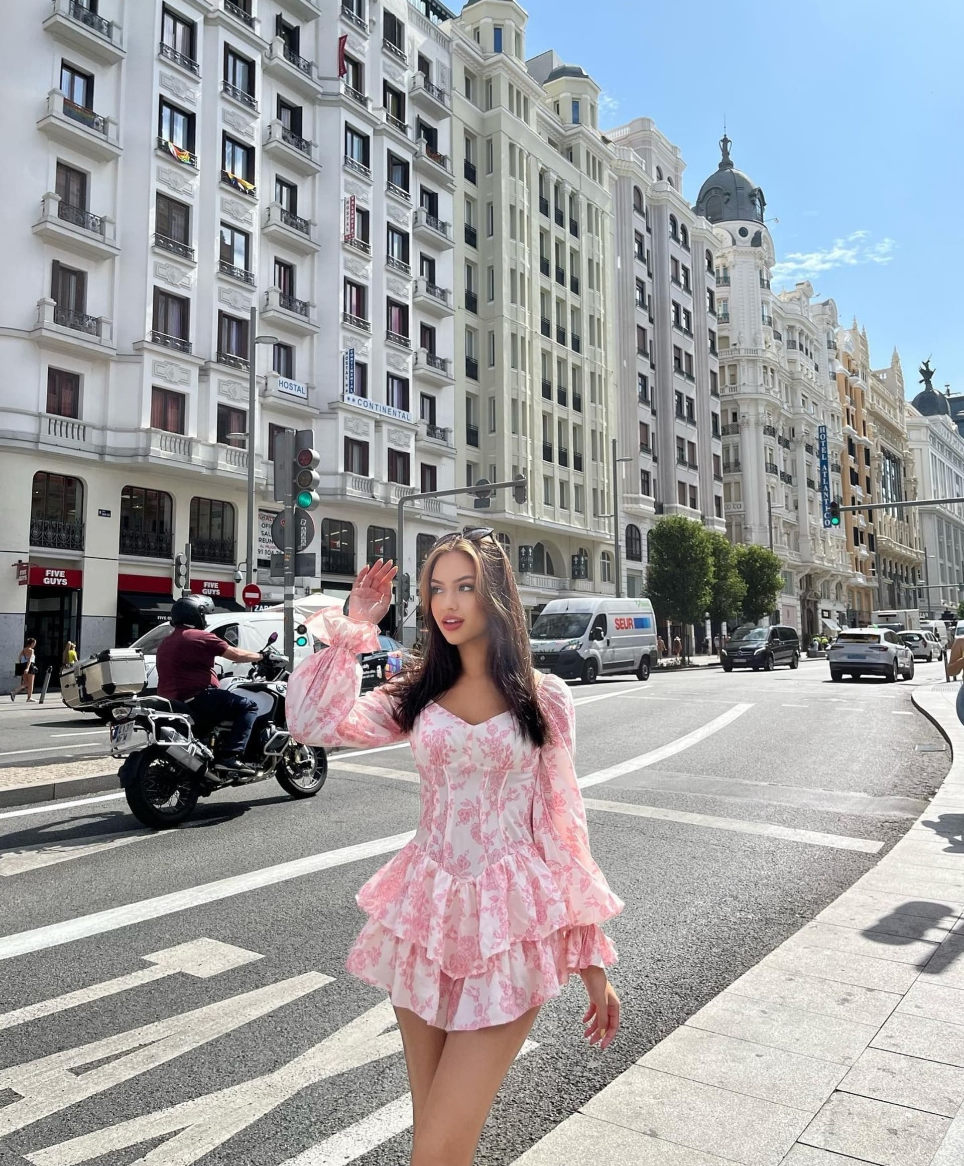 A woman in a pink floral dress waves from a city street corner near traffic.
