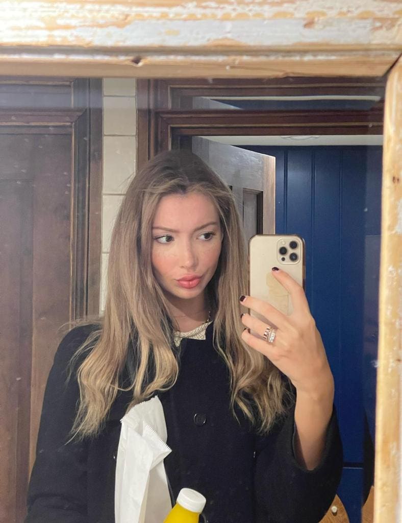 Mirror selfie of Clara in a dark coat holding a phone and paper bag in a room.