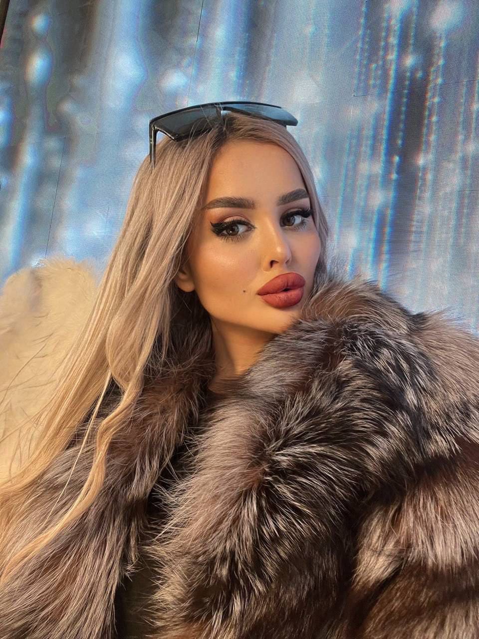 Astria con cabello rubio y gafas en la cabeza posa con abrigo de piel marrón frente a fondo azul brillante.
