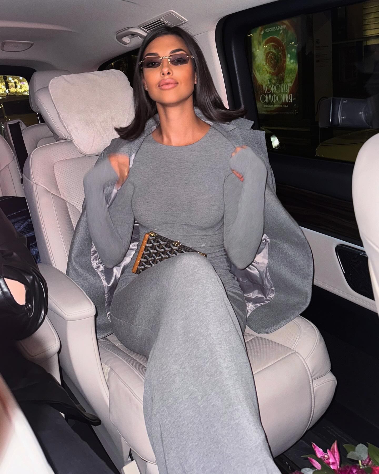 Mujer con cabello oscuro hasta los hombros y gafas pequeñas sentada en el asiento trasero de un coche con tapicería clara, llevando top gris ajustado y pantalones a juego.