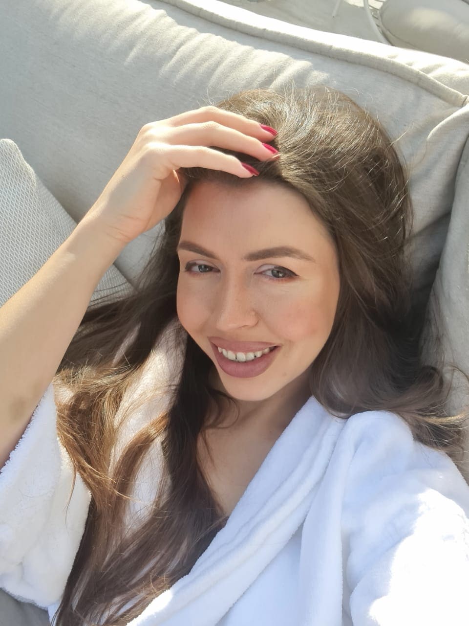 Mujer tumbada en cojín claro con bata blanca, sonriendo a la cámara mientras apoya su mano con uñas rojas sobre la frente.