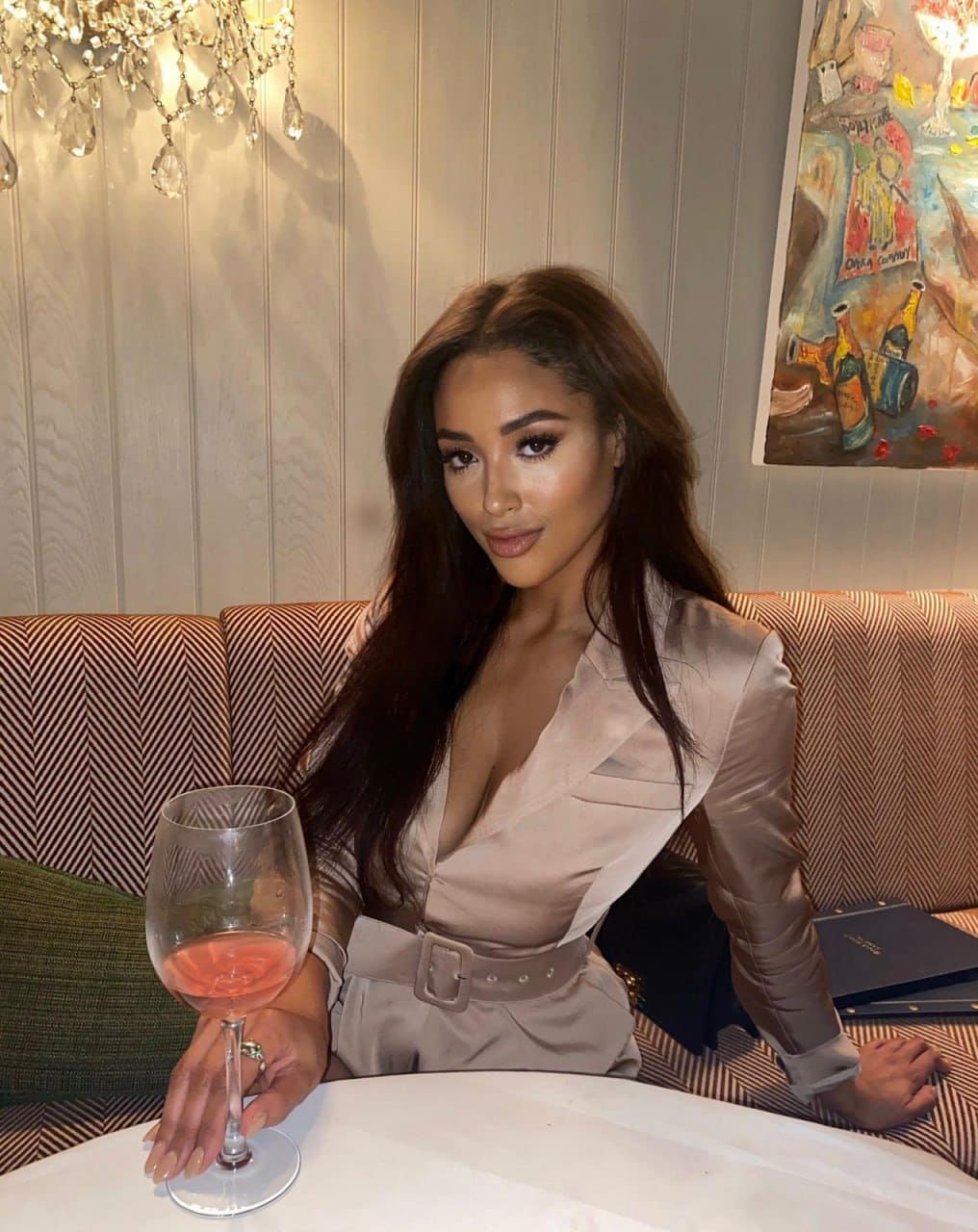 Donna con blazer satinato chiaro e cintura abbinata, seduta a un tavolo di ristorante con un calice di rosé.