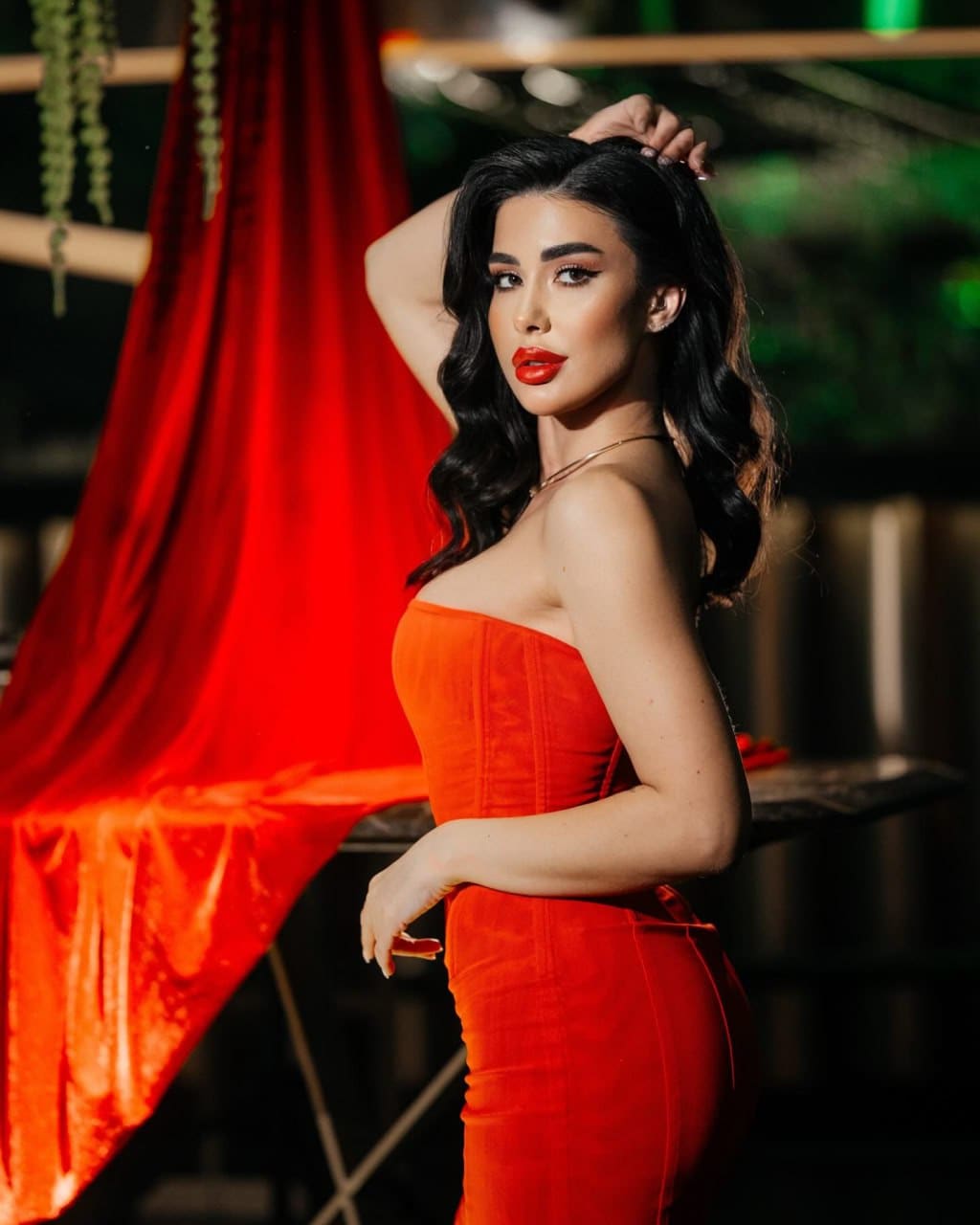Anna stands in profile in a strapless orange-red dress against red drapery and green light.
