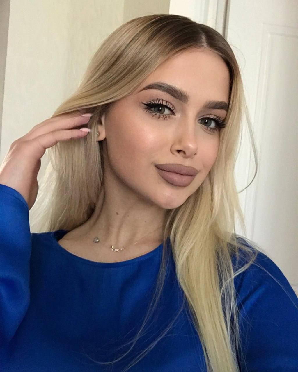A woman in a blue long-sleeved top raises one hand near her ear before a white door.