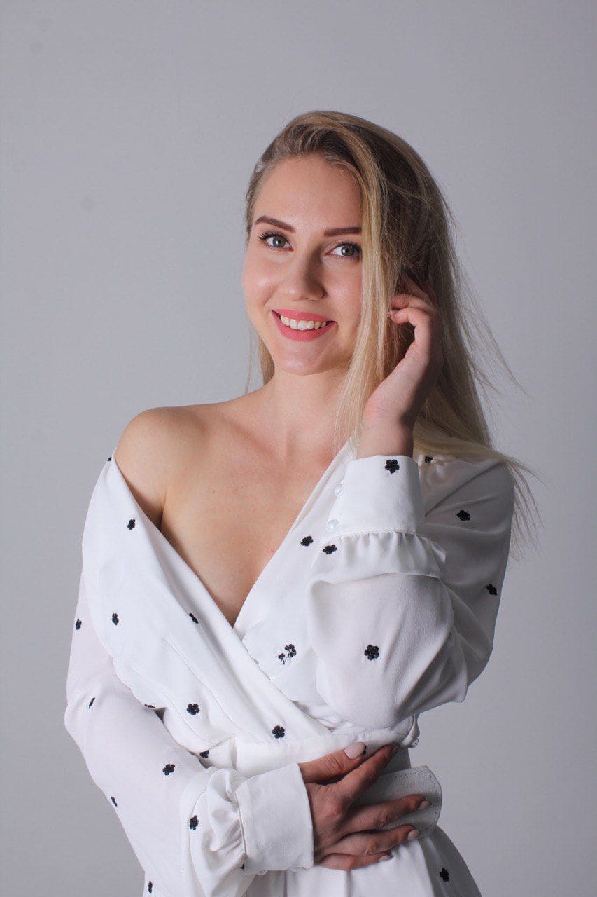 Bozhena de pie con fondo gris, blusa blanca bordada caída de un hombro y una mano en el cabello.